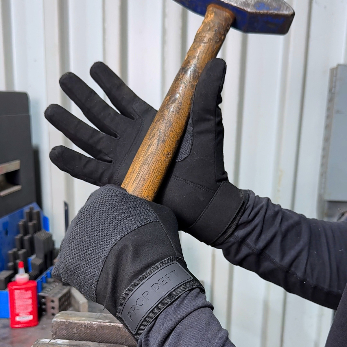 Black work gloves with textured grip panels and a visible 'PROP DEPT' label on the cuff, shown being worn while holding a wooden-handled hammer in a workshop setting.